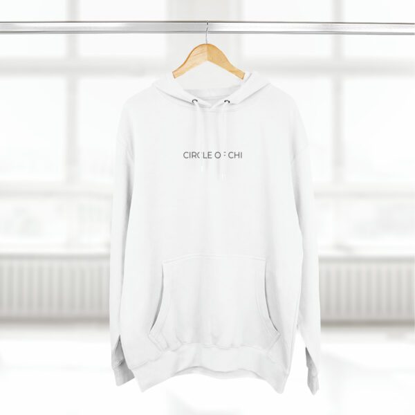 A white Three-Panel Fleece Hoodie with "CIRCLE:CHI" text on the front hangs on a wooden hanger against a bright window background.